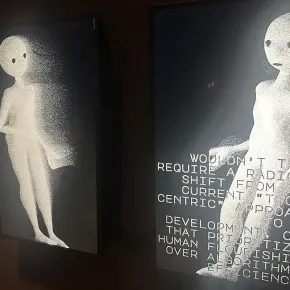 Two illuminated display screens in a darkened gallery space, each showing a grainy, stippled humanoid figure against a dark background. The right screen features overlaid text that reads: "Wouldn't that require a radical shift from our current 'tool-centric' approach to AI development, one that prioritizes human flourishing over algorithmic efficiency?"