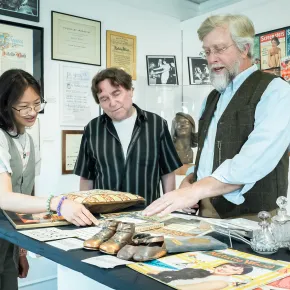 Teresa with two men reviewing what looks like Natalie Wood memorabilia — bronzed baby shoes, vintage magazines, cut-out doll books — in an exhibit or archive room. 