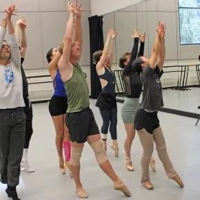 Dance students in a studio reach their arms overhead in unison during a ballet class, wearing rehearsal clothes and pointe shoes, with mirrors and barres lining the walls behind them.
