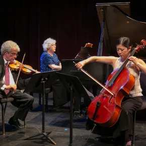 Faculty musicians perform on a darkened stage, with Haroutune Bedelian playing violin on the left, Lorna Griffitt at a grand piano in the center, and Sarah Koo Freeman playing cello on the right.