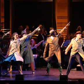 Performers in trench coats and fedoras point upward in unison onstage in a production choreographed by Andrew Palermo, with an orchestra and chorus seated behind them.