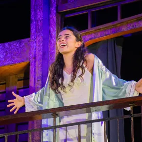 Lourdes Castillo as Juliet, arms outstretched and smiling on a balcony in a pale nightgown and robe, in New Swan Shakespeare Festival's Romeo & Juliet.