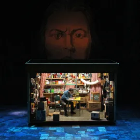 Production photo from Mother Russia, a small lit shop set cluttered with boxes and shelves of goods sits onstage as a man writes at a desk, beneath a giant painted face looming in the darkness above