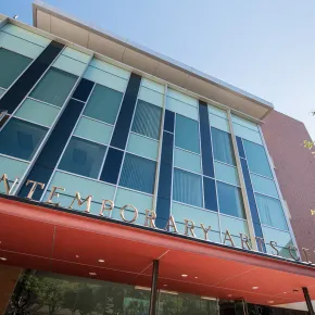 A low-angle view of the Contemporary Arts Center, with bronze letters spelling its name along a red-orange entrance canopy, a facade of teal and navy glass panels and a red brick wall against a clear blue sky.