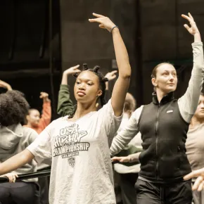 Dance students rehearse choreography in a studio, extending their arms in coordinated poses.