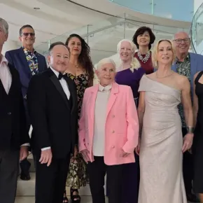 Nine OC Arts Awards honorees in formal attire pose together on a staircase with glass railings in a bright, modern lobby. A grand piano is visible behind them.