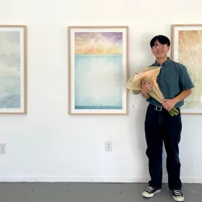 An artist holding a bouquet of flowers smiles beside three framed works on a white gallery wall. The works are soft, layered abstractions in blue, gold and green that suggest water and sky.