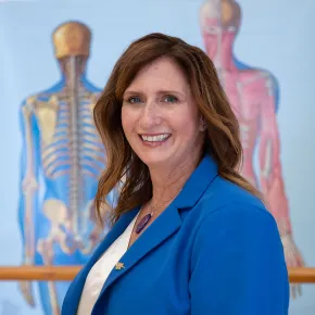 Dr. Kelli Sharp smiles at the camera, wearing a bright blue blazer over a white blouse. Anatomical illustrations of the human skeletal and muscular systems are displayed on the wall behind her.