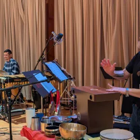 Camilo Zamudio plays marimba behind a drum kit as co-leader Wilfrido Terrazas gestures beside a cajón and a table of singing bowls and frame drums during a rehearsal in a wood-paneled studio.
