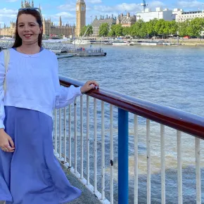 Emily Parise, in a white sweater and periwinkle dress, smiles beside a railing on the River Thames, with Big Ben and the Houses of Parliament in the background.