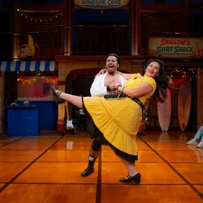 On a sunlit wooden stage dressed as a beachside boardwalk, with signs for “Windsor Cove” and “Shallow’s Surf Shack,” a laughing man in an open white shirt dips a smiling woman in a yellow polka-dot top and full yellow skirt as she kicks one leg high. Audience members seated around the stage edge look on and smile.
