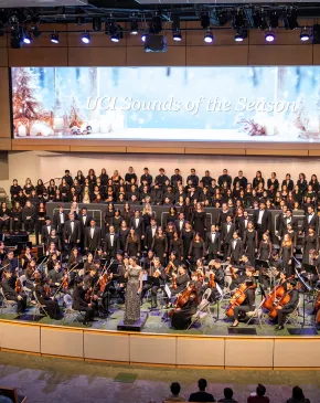 A full orchestra and choir perform together in a large concert hall, filling the stage with musicians and singers. A conductor stands at the center leading the ensemble, while a seasonal backdrop reading “UCI Sounds of the Season” spans the stage above them.
