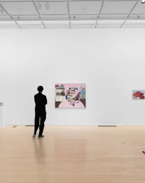 A visitor stands in a contemporary art gallery, viewing wall-mounted artworks and a hanging mixed-media installation.
