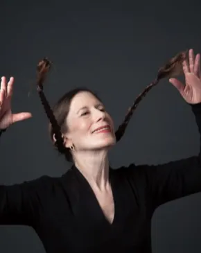 A performer poses expressively against a dark background, raising her hands as her braided hair extends outward.