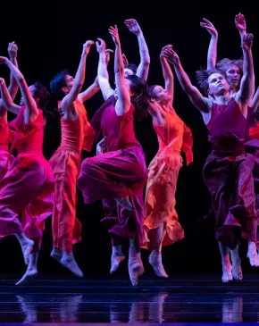 A group of dancers leap and move together on stage in flowing red costumes during a contemporary dance performance.