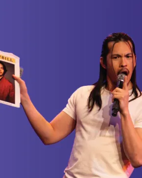 Performer onstage speaks into a microphone while holding up a Playbill