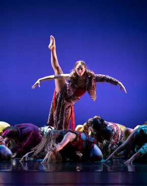 A dancer balances with one leg extended overhead while an ensemble kneels around them under blue stage lighting.