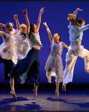 Five dancers leap simultaneously on a dimly lit stage, arms raised, wearing flowing white and navy garments against a deep blue backdrop.