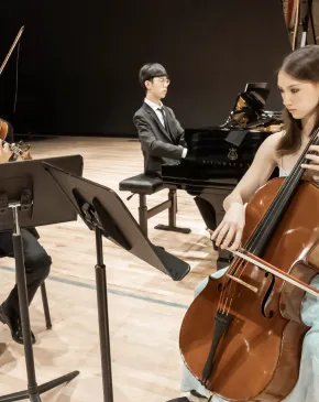 Three musicians perform on a concert stage, with a violinist and cellist reading from music stands and a pianist seated at a grand piano behind them.