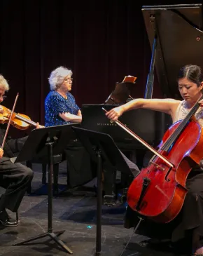 Faculty musicians perform on a darkened stage, with Haroutune Bedelian playing violin on the left, Lorna Griffitt at a grand piano in the center, and Sarah Koo Freeman playing cello on the right.