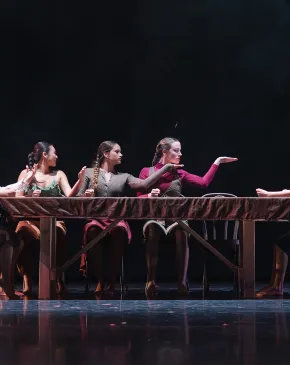 Dancers perform a theatrical piece seated along a draped table on a darkened stage, gesturing expressively toward one another.