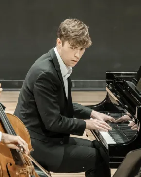 Two musicians in formal black attire perform together, one playing cello and the other seated at a Steinway grand piano.