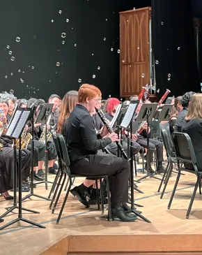 A large ensemble of musicians dressed in black performs woodwind and saxophone instruments on a concert stage with soap bubbles floating in the air above them.