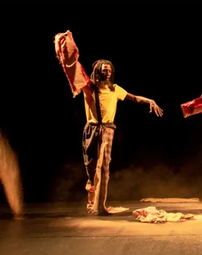 Three dancers perform on a dimly lit stage, each manipulating large pieces of red and earth-toned fabric with outstretched arms and dynamic movement.