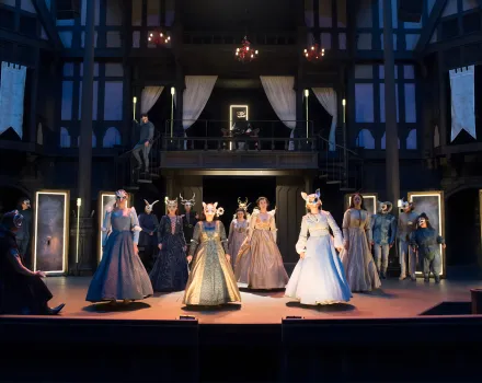 A large cast stands across a theater stage in elaborate period costumes and masks, arranged in formation beneath warm stage lighting, with a multi-level wooden set and balconies visible behind them.