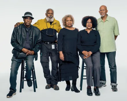 Five adults pose together for a studio portrait against a plain light background, arranged in a mix of seated and standing positions. They wear casual to semi-formal clothing in dark and muted tones, standing close with relaxed, composed expressions, conveying a sense of familiarity and shared presence.