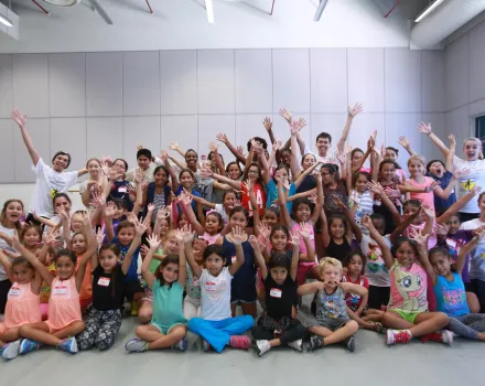 A large group of children and a few adults pose together indoors, seated and standing in rows. Many raise their hands and smile toward the camera. The room has light-colored walls and a high ceiling with exposed fixtures.