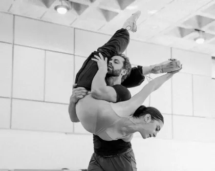 Two dancers perform an acrobatic lift in a bright studio, with one dancer held inverted and arched while the other supports them securely. The black-and-white image emphasizes strength, balance, and controlled movement.