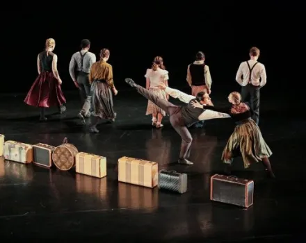 A group of performers moves across a dark stage with vintage-style costumes, while suitcases and trunks are arranged in a line along the floor. One performer balances in a lifted, extended pose as others walk or turn away, creating a layered stage composition.