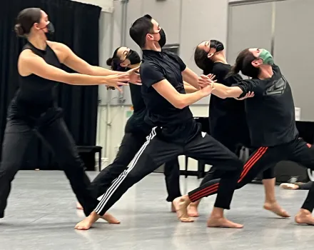 Group of dancers in a studio wearing black rehearsal clothing and face masks, performing a synchronized movement while leaning and reaching together.
