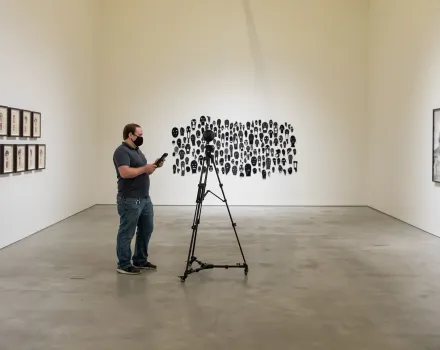 Person standing in a gallery beside a tripod-mounted camera, facing a wall installation composed of many small black silhouette portraits, with additional framed works displayed on the surrounding walls.
