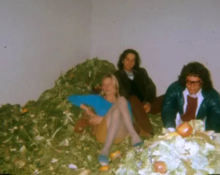 Three people seated on the floor beside a large pile of organic debris or compost material, including leaves and discarded food items, inside a plain room.
