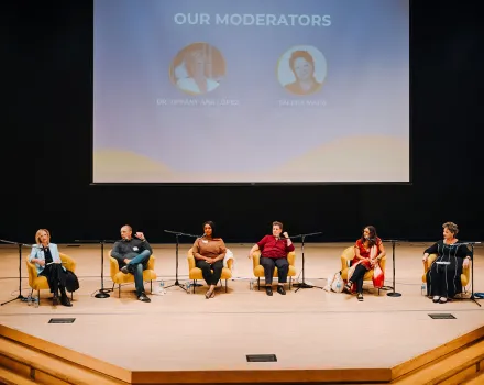 A panel of six speakers sits onstage in armchairs with microphones, participating in a discussion. A large screen behind them displays the heading “Our Moderators” along with two profile images, while the audience area is out of view.