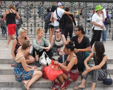 A group of people sits together on stone steps, talking and resting in front of a decorative metal fence. Some hold bags and personal items, including a bright red tote placed in the center. Other pedestrians move in the background.