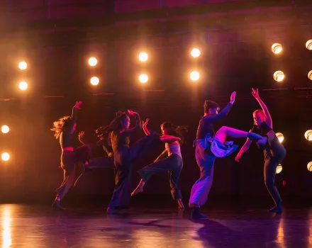 A group of dancers performs onstage under bright amber stage lights. They move dynamically in pairs and small clusters, with lifted legs and raised arms silhouetted against the glowing backdrop.