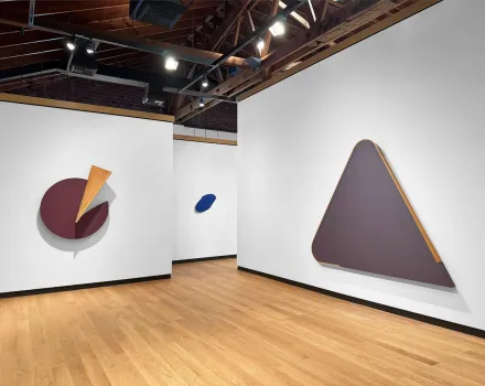 A gallery room with white walls and a light wood floor displays large geometric wall-mounted artworks. On the left wall, a circular maroon shape is partially overlaid with a yellow triangular form. In the center background, a small blue oval piece is mounted. On the right wall, a large triangular form in muted purple with a narrow wooden edge detail is displayed. Overhead track lighting illuminates the space, and exposed wooden beams are visible on the ceiling.