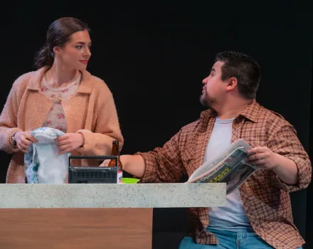 Two actors stand behind a kitchen-style counter on a stage set. One person in a tan jacket holds a dish towel and looks to the side at the other actor, who wears a brown plaid shirt over a white T-shirt and holds a folded newspaper. A small radio and kitchen items sit on the countertop, and the dark stage background isolates the pair under warm lighting.