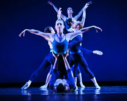 A group of dancers performs on a stage under deep blue lighting. One dancer stands at the front with arms extended outward, while others are positioned closely behind and around her, creating layered, symmetrical shapes with their raised arms and legs. Another dancer lies on the floor at the base of the formation, adding to the dramatic, sculptural composition.