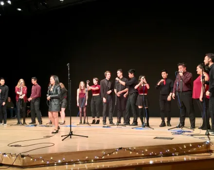 A group of performers stands across a stage in a theater, dressed mostly in black with red accents. Several hold microphones while others stand near mic stands, appearing to sing or prepare to perform. String lights line the front edge of the stage, and the background is a dark auditorium setting with overhead stage lighting.
