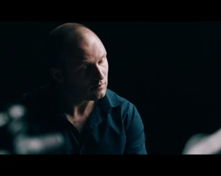 Moody, low-light portrait of a bald man in a dark shirt looking downward in a dim setting, with soft highlights on his face and a blurred foreground element suggesting a table or instrument.