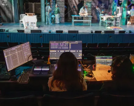View from a theater control booth showing technicians at lighting and sound consoles in the foreground, with actors rehearsing on a lit stage in the background.