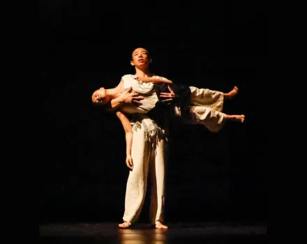 Two contemporary dancers perform a partnered lift on a dark stage, with one dancer standing and supporting another held horizontally across their torso under focused theatrical lighting.