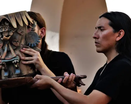 Fabricio Cavero, with long dark hair and wearing a black shirt, holds a large ceremonial clay vessel adorned with intricate pre-Columbian sculptural figures, including a skeletal deity and stylized hands, during a live performance. A second performer with long hair, partially obscured behind the vessel, helps support it. The piece is richly detailed with earthy brown and gray tones, and the background suggests an outdoor or open architectural setting with soft, bright light.