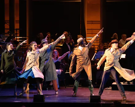 Performers in trench coats and fedoras point upward in unison onstage in a production choreographed by Andrew Palermo, with an orchestra and chorus seated behind them.