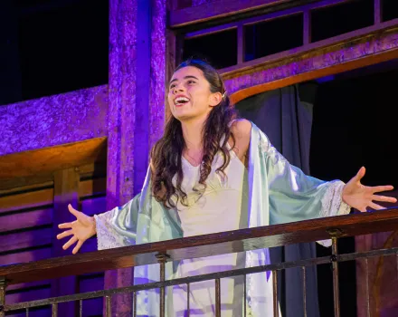 Lourdes Castillo as Juliet, arms outstretched and smiling on a balcony in a pale nightgown and robe, in New Swan Shakespeare Festival's Romeo & Juliet.