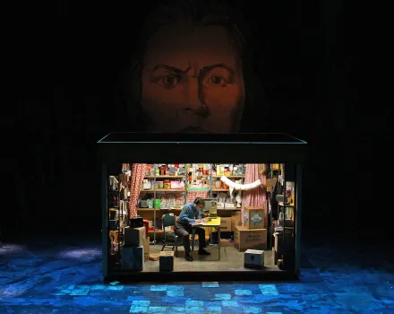 Production photo from Mother Russia, a small lit shop set cluttered with boxes and shelves of goods sits onstage as a man writes at a desk, beneath a giant painted face looming in the darkness above